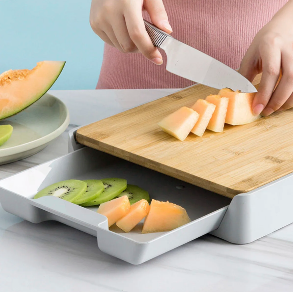 Person slicing melon on a wooden cutting board with a knife, using a white tray below to catch the pieces.