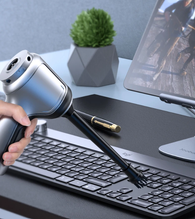 Hand using a keyboard cleaning tool on a desk with a laptop and plant in the background